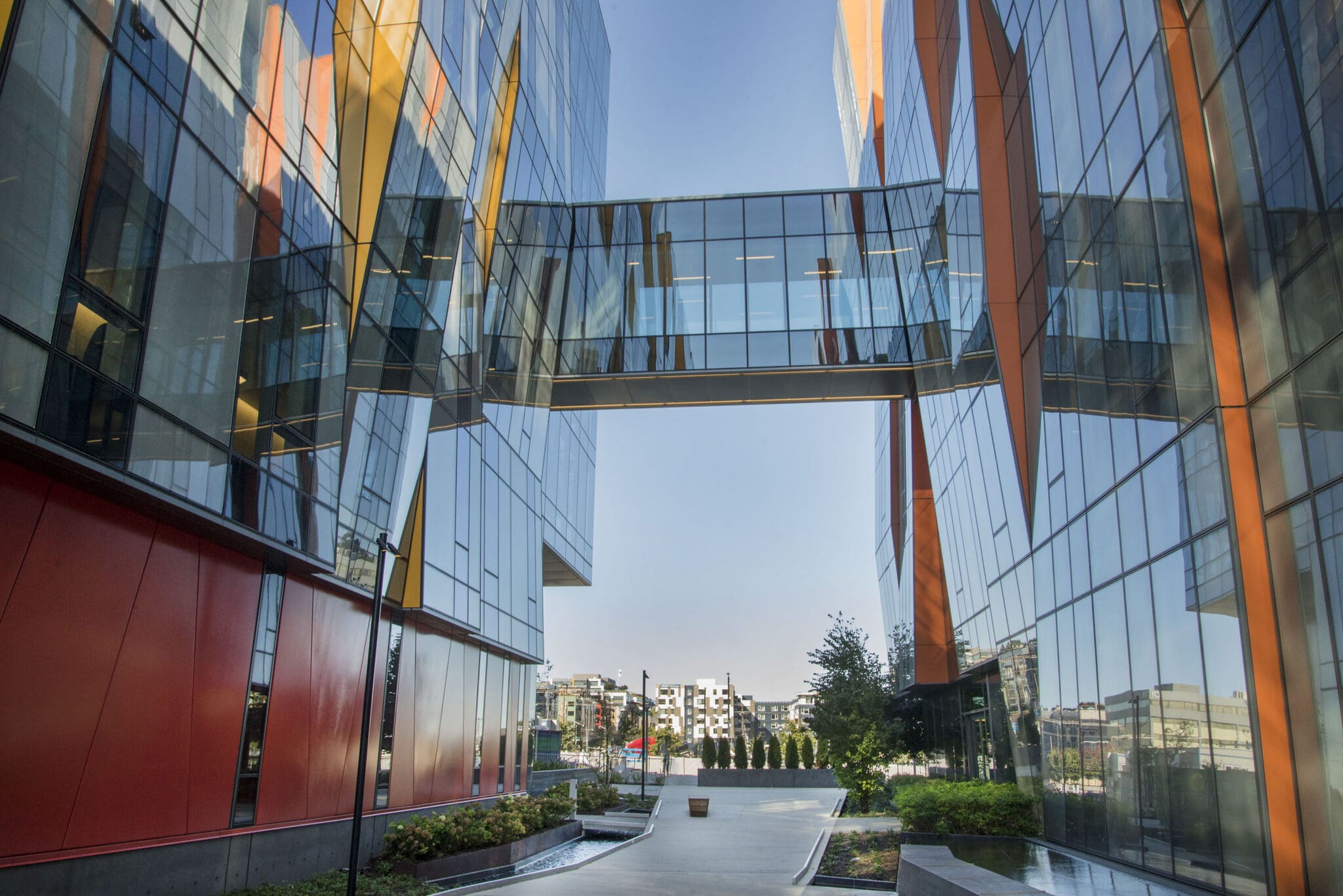 A modern building with glass walls and orange accents features a glass skybridge connecting two sections above a landscaped walkway with shrubs and trees.