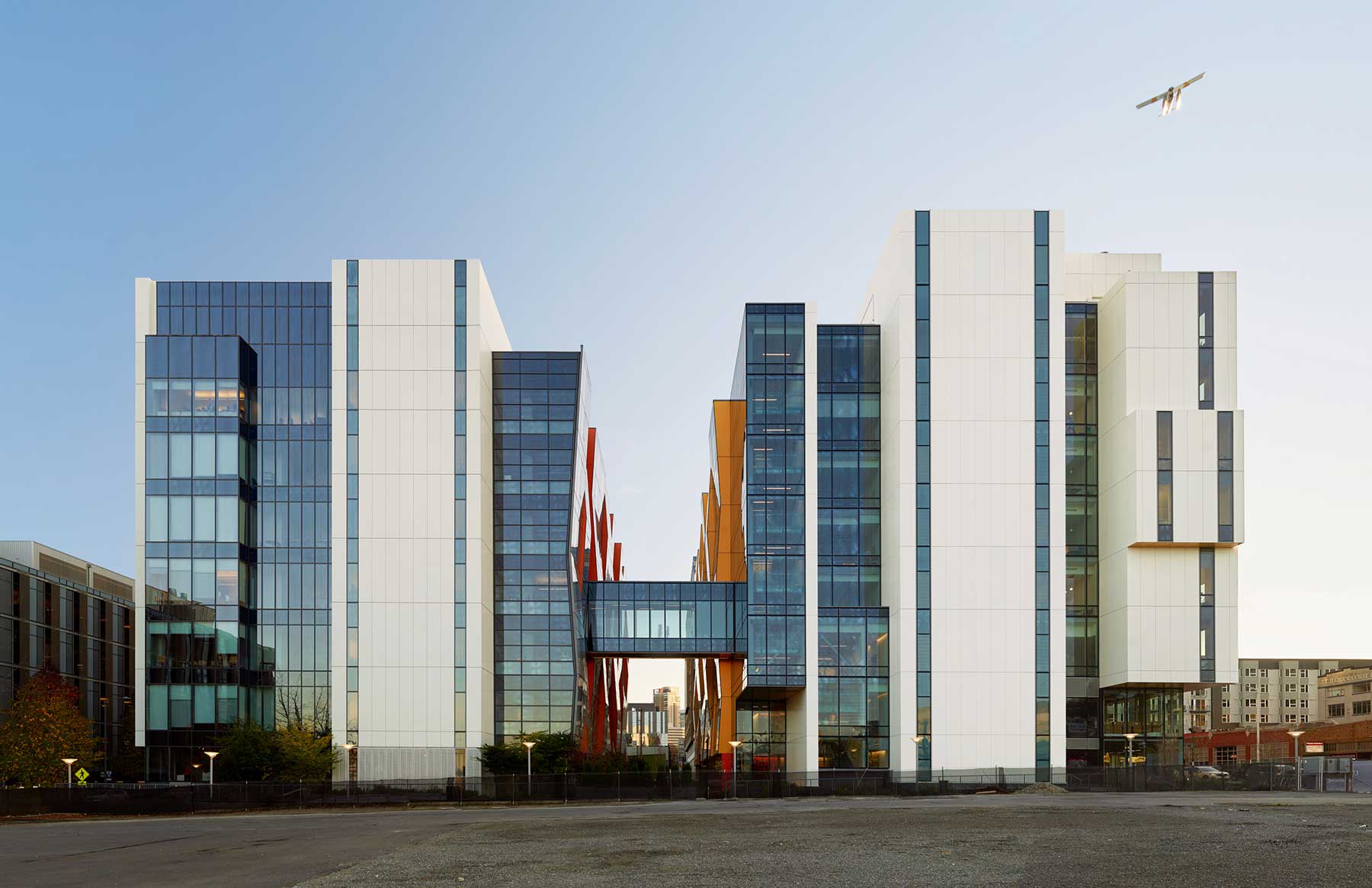 A modern glass and white-paneled building with a skybridge connects two towers; red architectural details are visible inside. An airplane flies in the clear blue sky above.