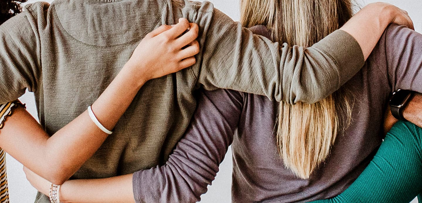 Three people are standing side by side with their arms around each others shoulders, showing unity and support. The image is cropped to show only their backs and arms.