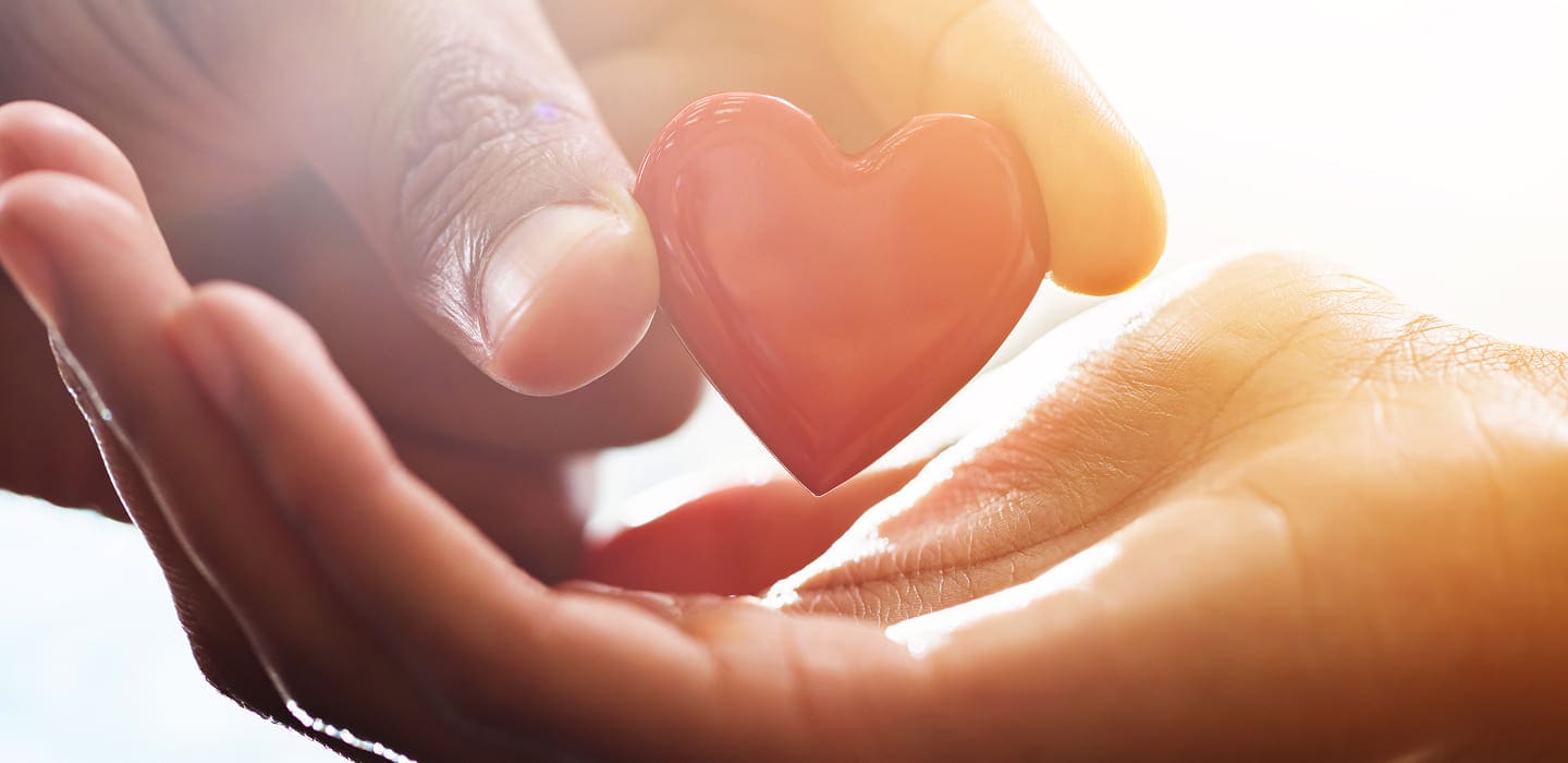 A close-up of one hand gently placing a small red heart-shaped object into another open hand, symbolizing care, love, or support, with a soft, bright background.