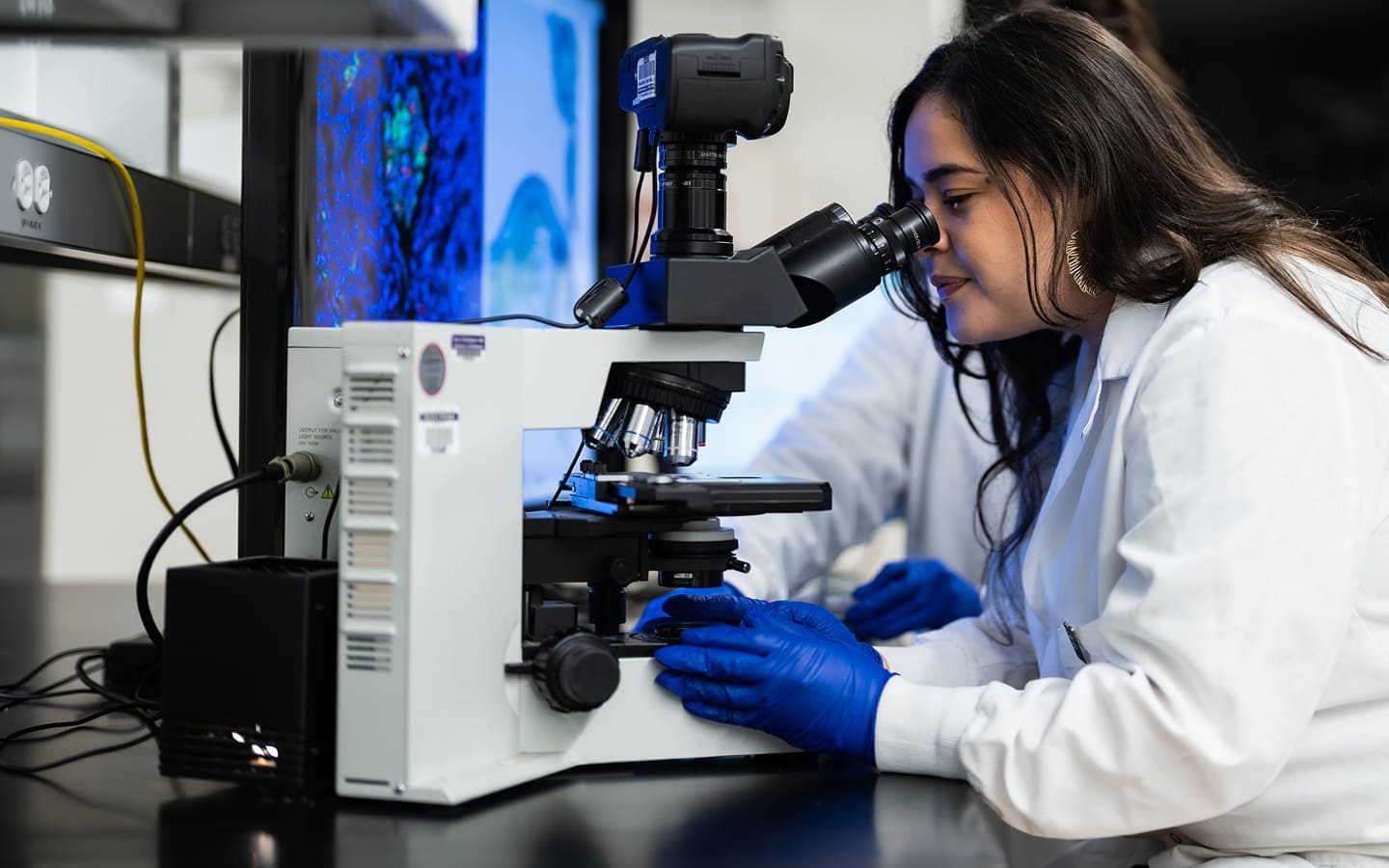 A scientist wearing a white lab coat and blue gloves looks into a microscope in a laboratory, with a computer screen displaying scientific images in the background.