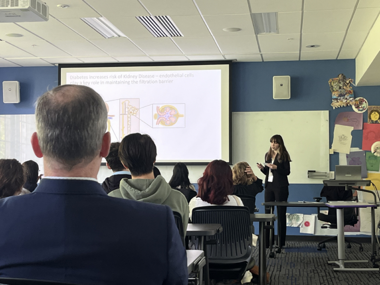 A woman presents a medical slide about kidney disease to a seated audience in a classroom, with colorful drawings on the wall and people listening attentively.