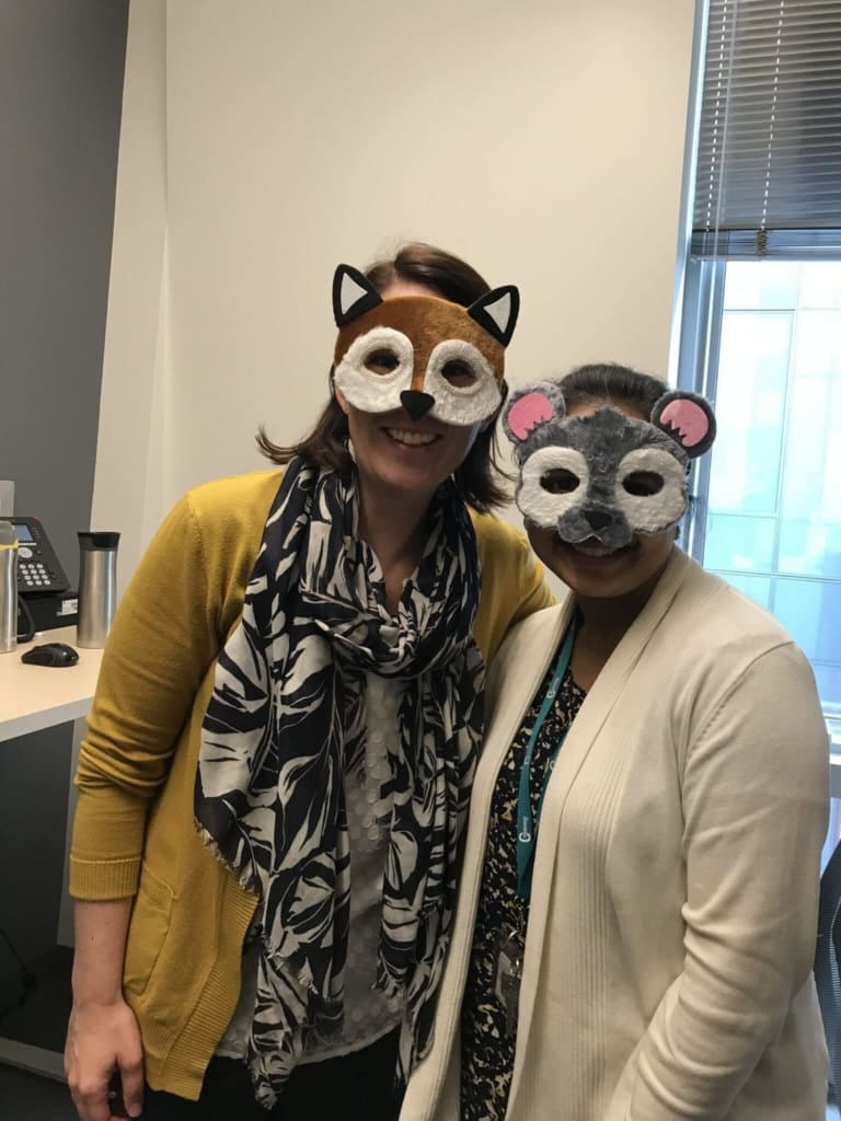 Two people standing side by side in an office, smiling and wearing animal masks—one resembling a fox and the other resembling a mouse. Both are dressed in casual work attire.