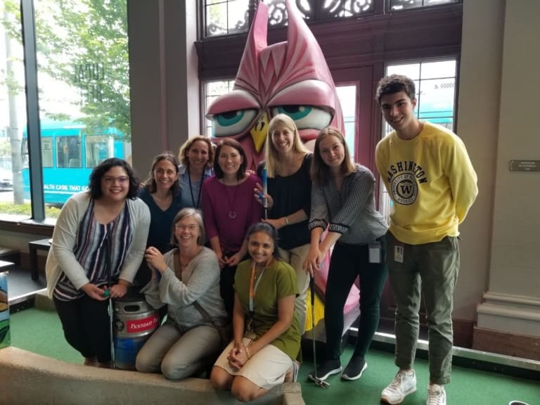 A group of nine smiling people poses indoors in front of a large pink owl statue. Most are standing, two are kneeling, and one person is wearing a yellow Washington sweatshirt. A window and greenery are visible in the background.