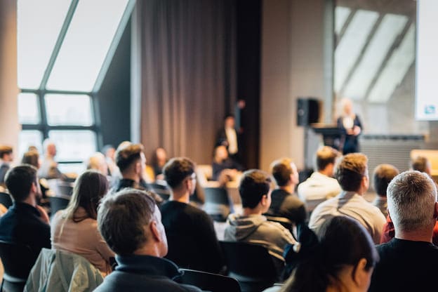 A group of people sit facing forward in a conference room, listening to a speaker presenting at the front near a screen. The room is bright with large windows and modern decor.
