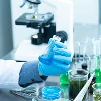 A gloved hand holds a flask containing blue liquid in a laboratory setting, with various glassware and a microscope visible on the table.
