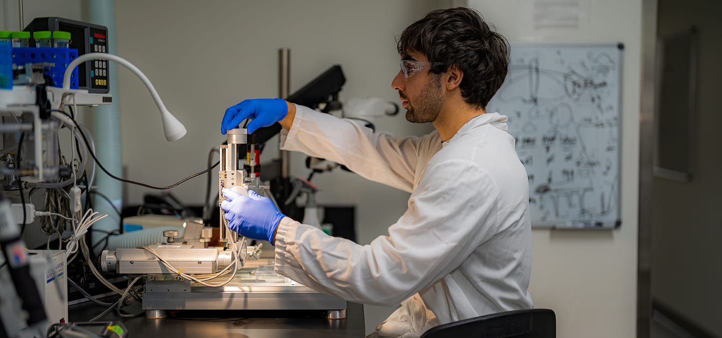 A scientist in a white lab coat and blue gloves works with laboratory equipment at a workstation, adjusting a device. Scientific diagrams and instruments are visible in the background.