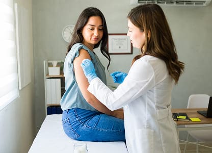 A healthcare professional wearing gloves administers a vaccine to a young woman sitting on an exam table in a medical office. The woman looks at her arm while receiving the injection.
