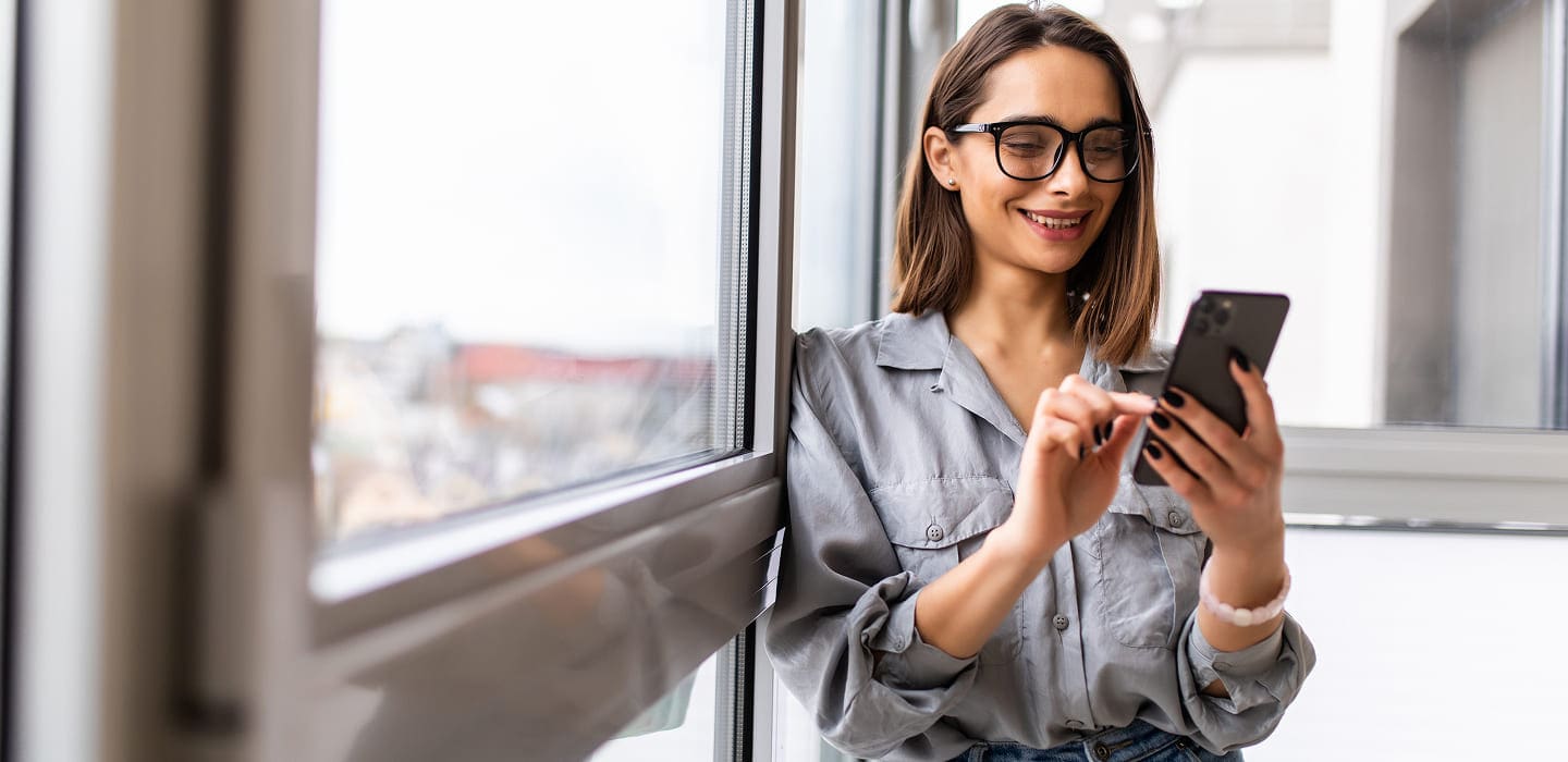 A smiling woman wearing glasses and a gray shirt stands by a window, looking at her smartphone and using it with one hand.