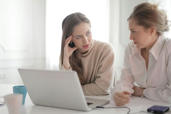 Two women sit at a desk with a laptop in front of them, engaged in a serious conversation. One woman holds a phone to her ear while listening intently; the other looks at her with concern.