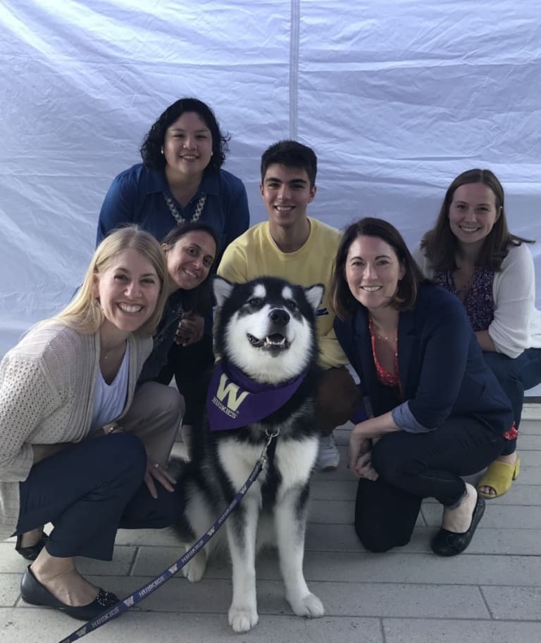 Six smiling people gather around a Siberian Husky wearing a purple bandana with a gold W on it, in front of a white backdrop outdoors.