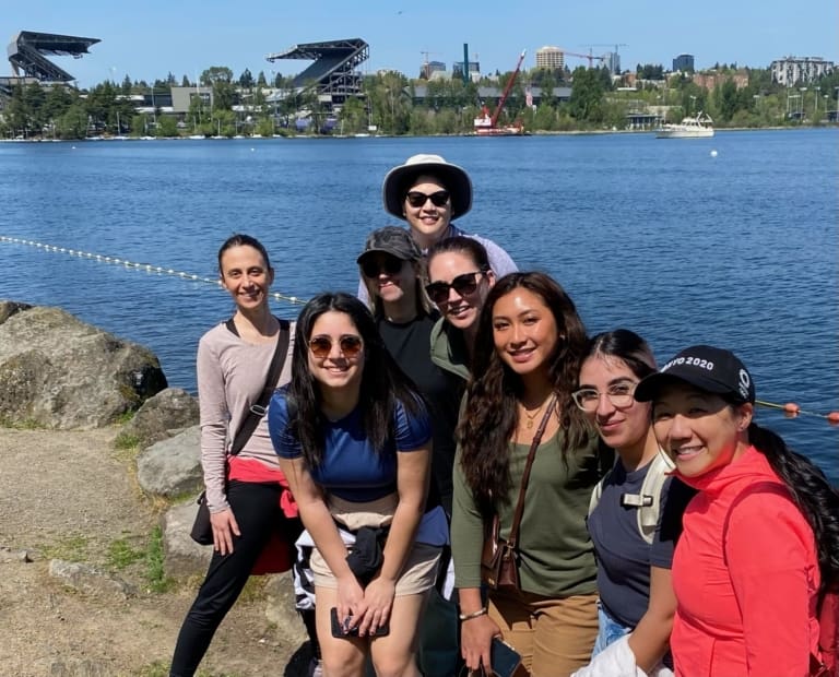 A group of eight smiling people pose together by a lakeshore on a sunny day, with water, trees, and stadiums visible in the background. Some wear sunglasses and hats.