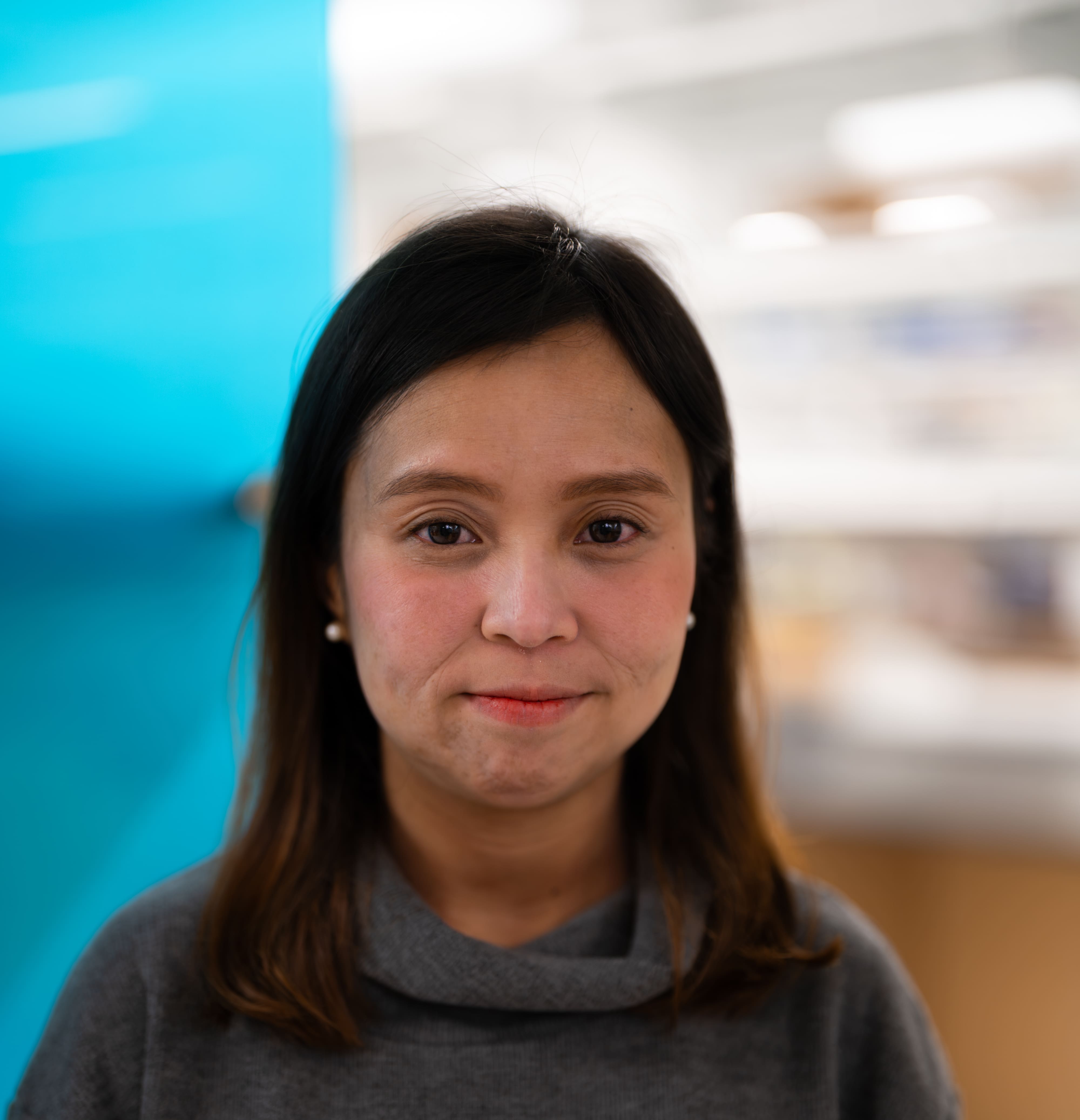 A woman with straight brown hair and a gray sweater smiles softly at the camera, standing indoors with a blurred blue and white background.