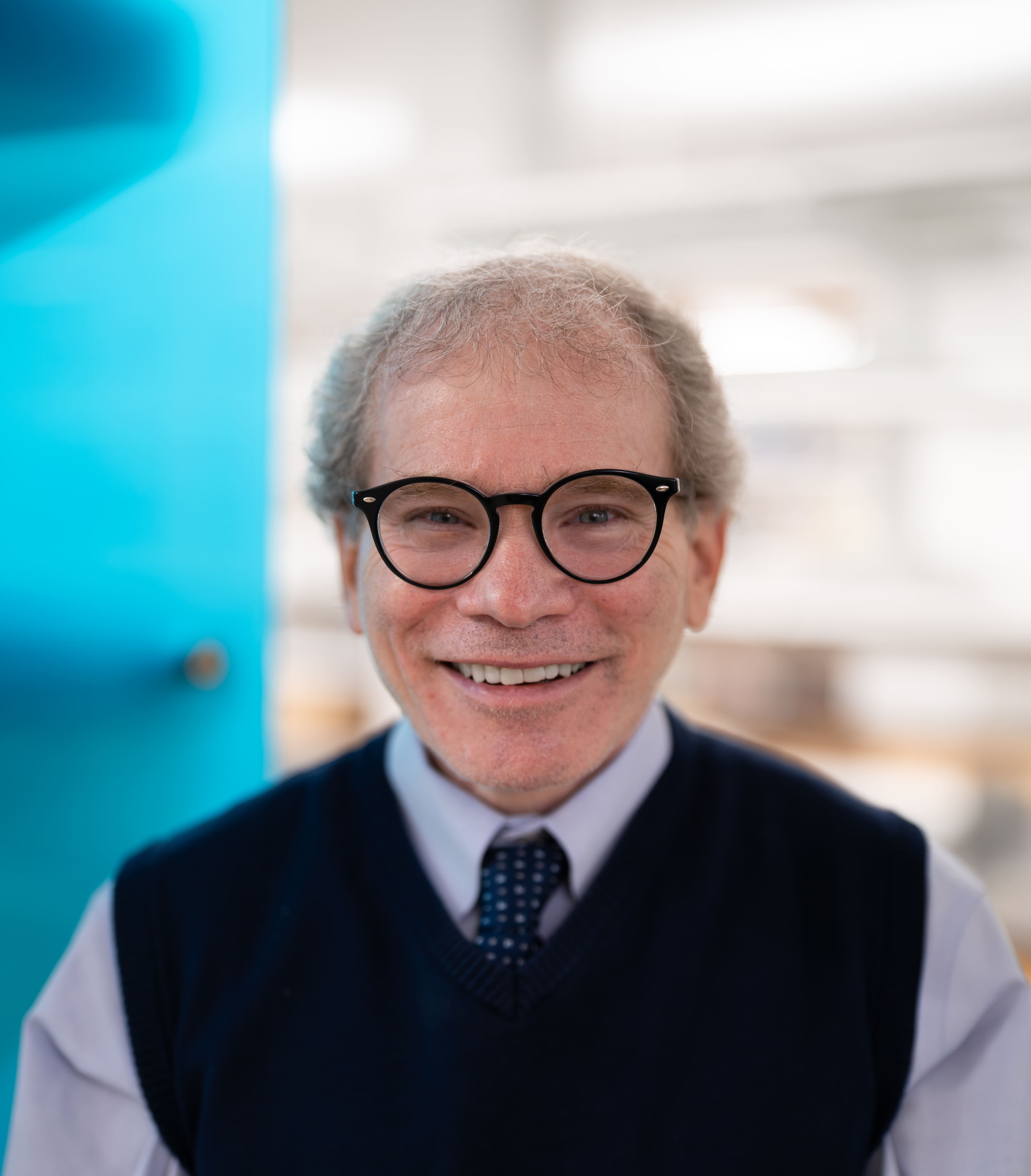 A smiling older man with gray hair, round black glasses, and a navy sweater vest over a collared shirt and tie, standing in a bright, blurred indoor setting.