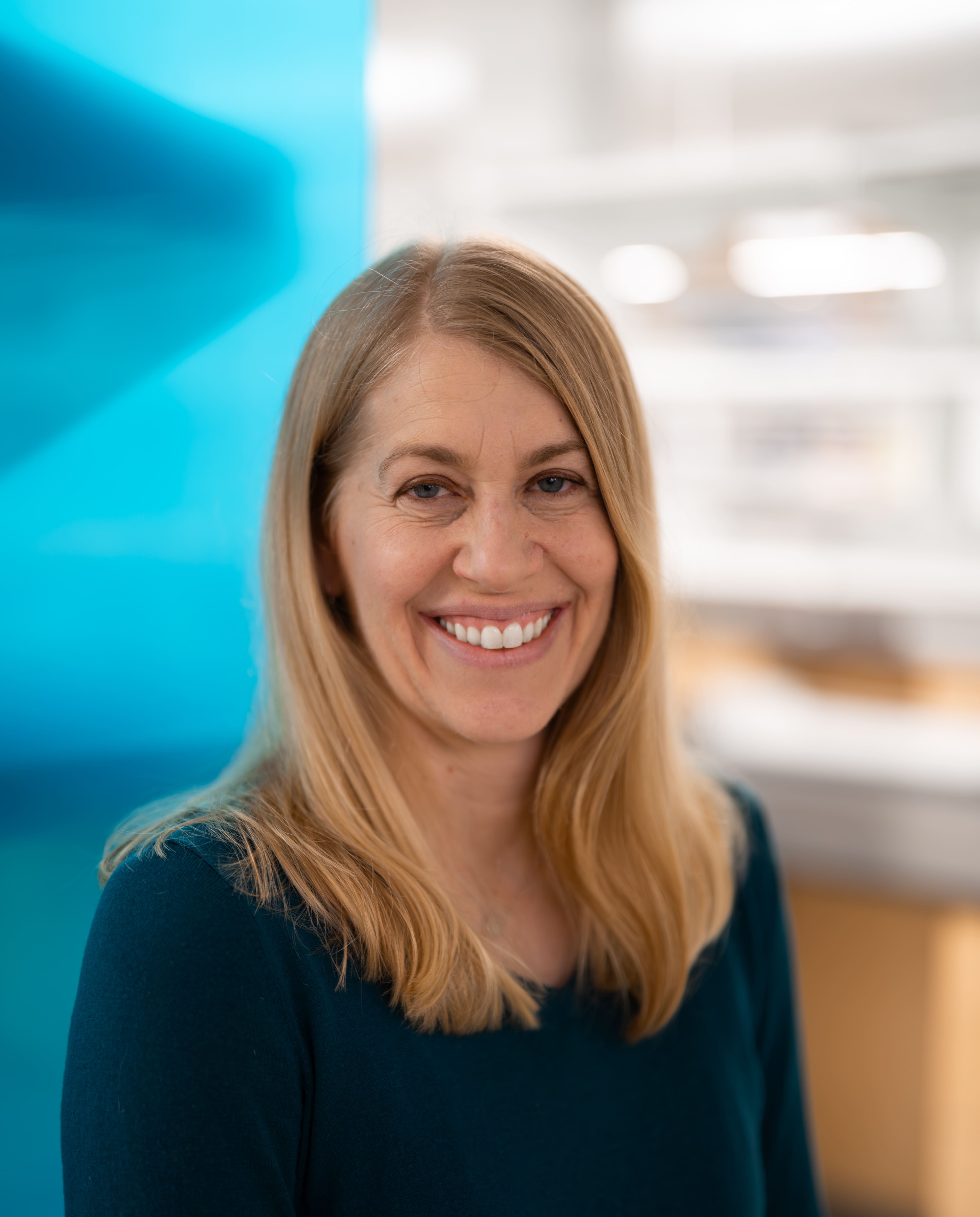 A woman with long blonde hair and a teal sweater smiles at the camera. The background is softly blurred with light and blue tones, suggesting an indoor setting.