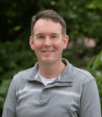 A man with short brown hair, wearing a light gray pullover with white stripes, smiles while standing outdoors in front of blurred green foliage.
