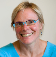 A smiling person with short blonde hair, wearing blue-framed glasses, turquoise earrings, and a light blue top, in front of a plain light background.