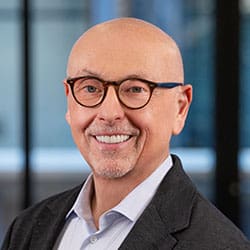 A smiling bald man with glasses and a goatee, wearing a dark blazer over a light-colored shirt, standing in front of a blurred office background.
