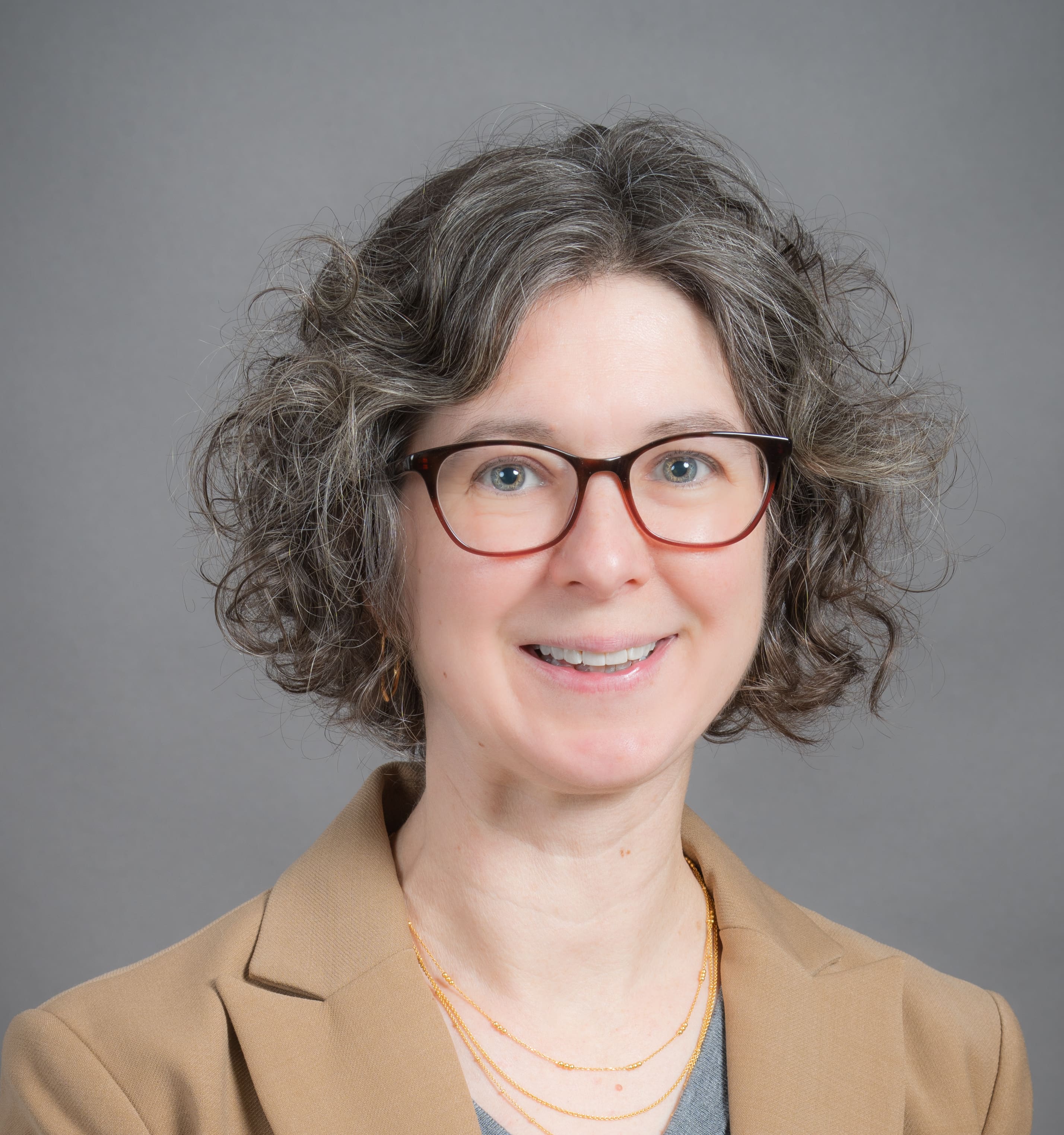 A woman with curly gray hair, wearing glasses, a tan blazer, and layered gold necklaces, smiles in front of a plain gray background.