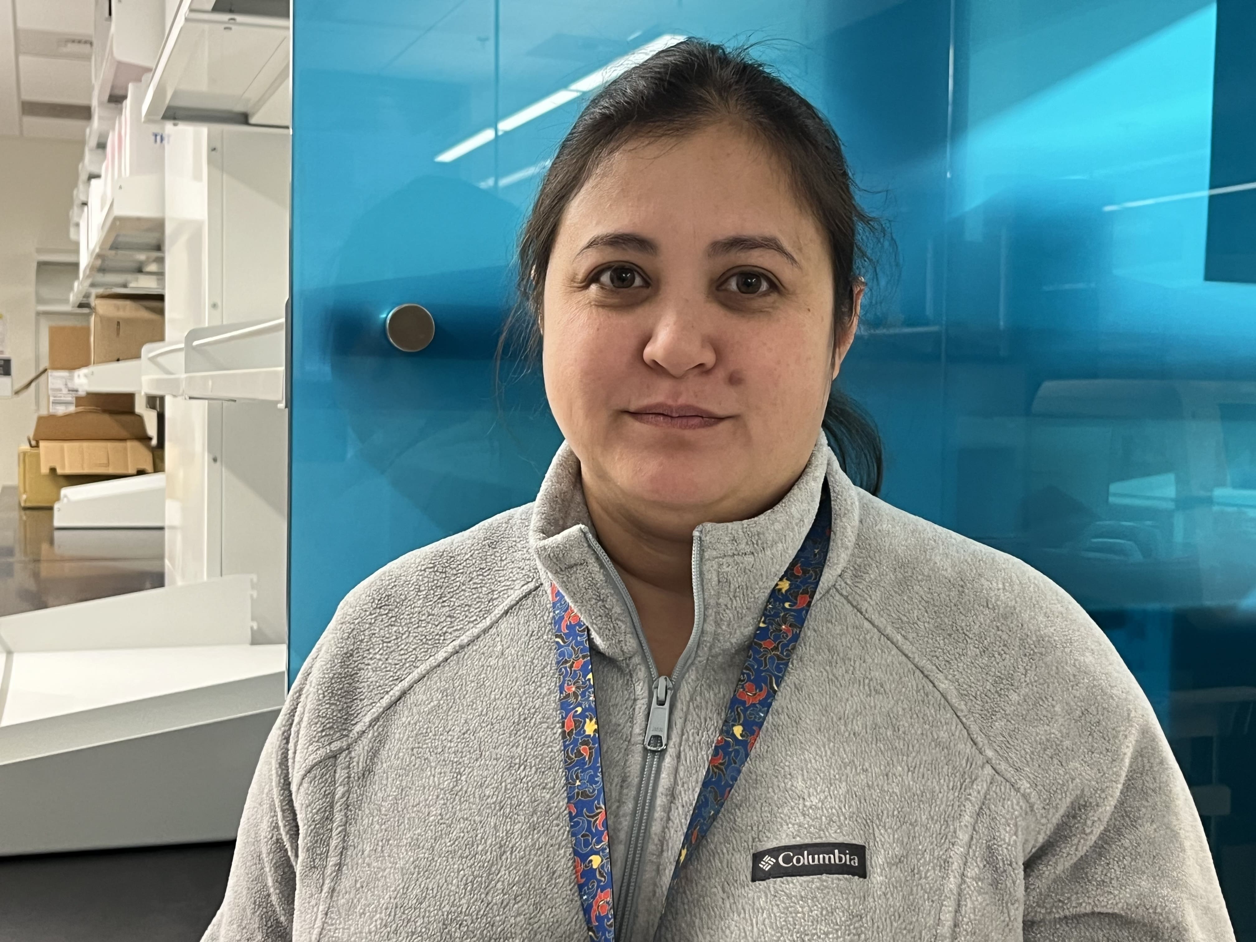 A woman wearing a light gray Columbia fleece and a patterned lanyard stands in a modern laboratory with blue cabinets and shelves in the background.