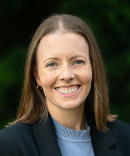 A woman with straight, light brown hair and blue eyes is smiling. She is wearing a black blazer over a light blue top and gold hoop earrings, with a blurred green background behind her.