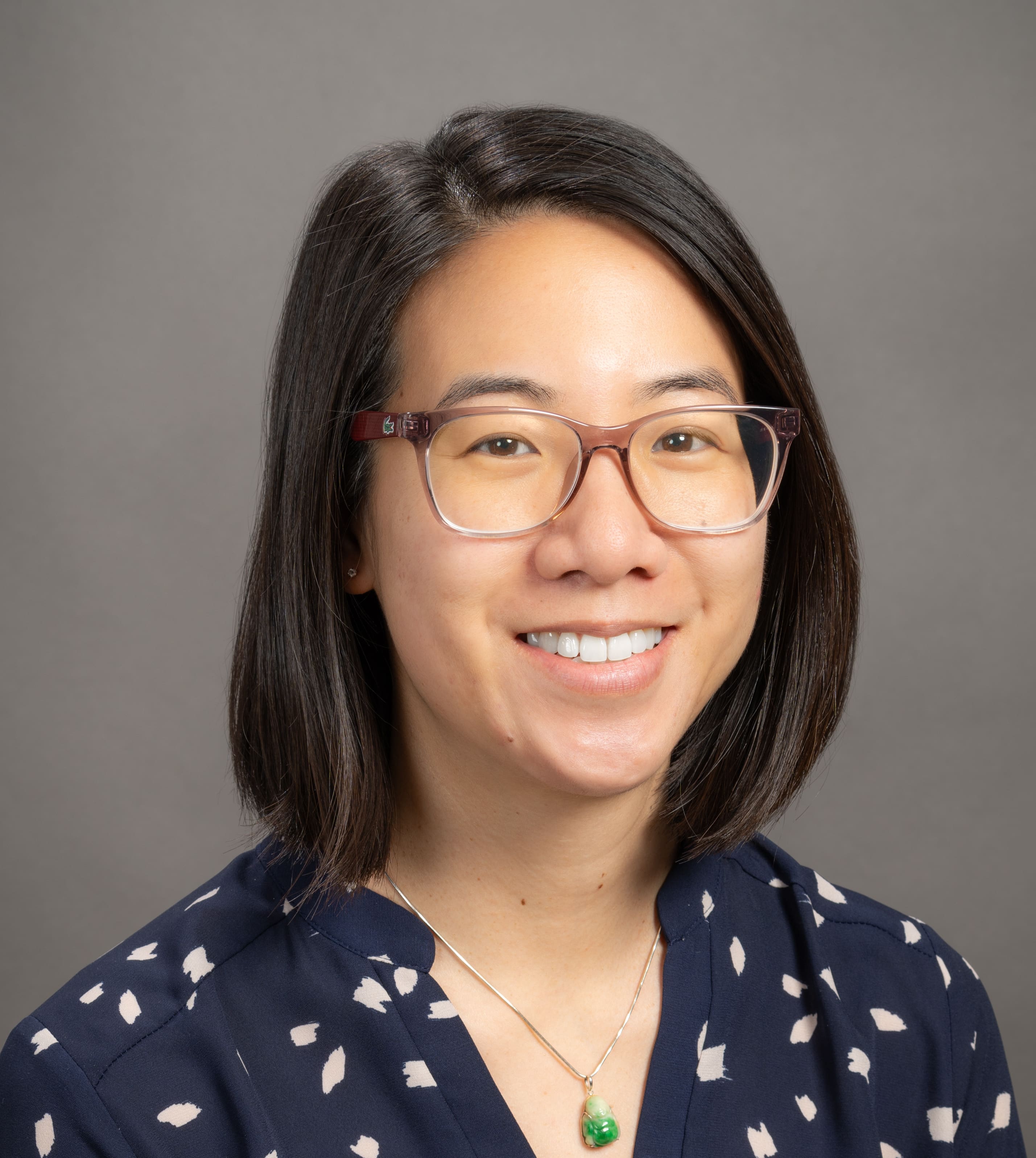 A woman with straight shoulder-length dark hair and glasses smiles at the camera. She is wearing a navy blouse with white patterns and a necklace with a green pendant, in front of a plain gray background.
