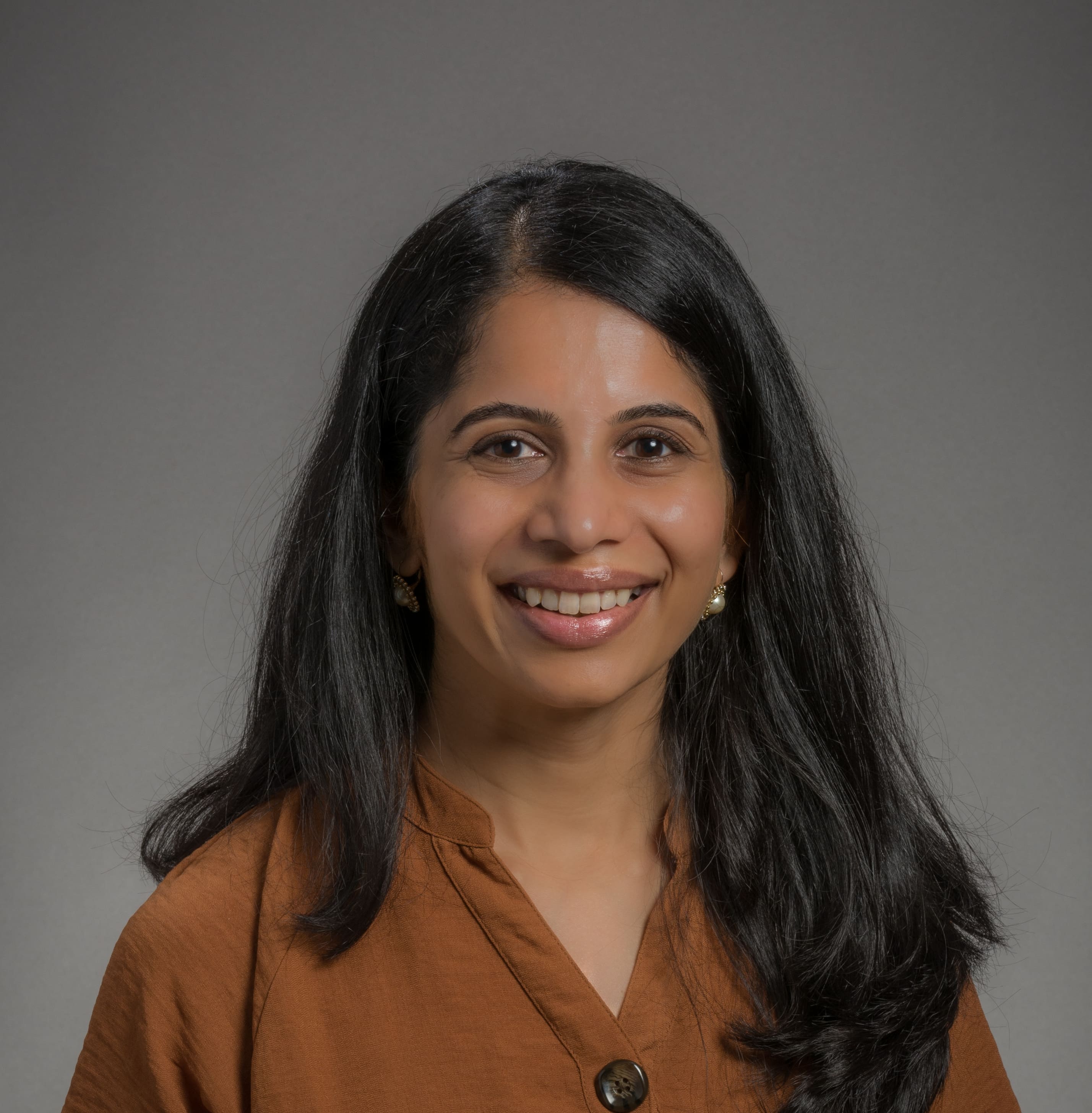A woman with long black hair is smiling at the camera. She is wearing a brown button-up top and small earrings, posed against a plain gray background.