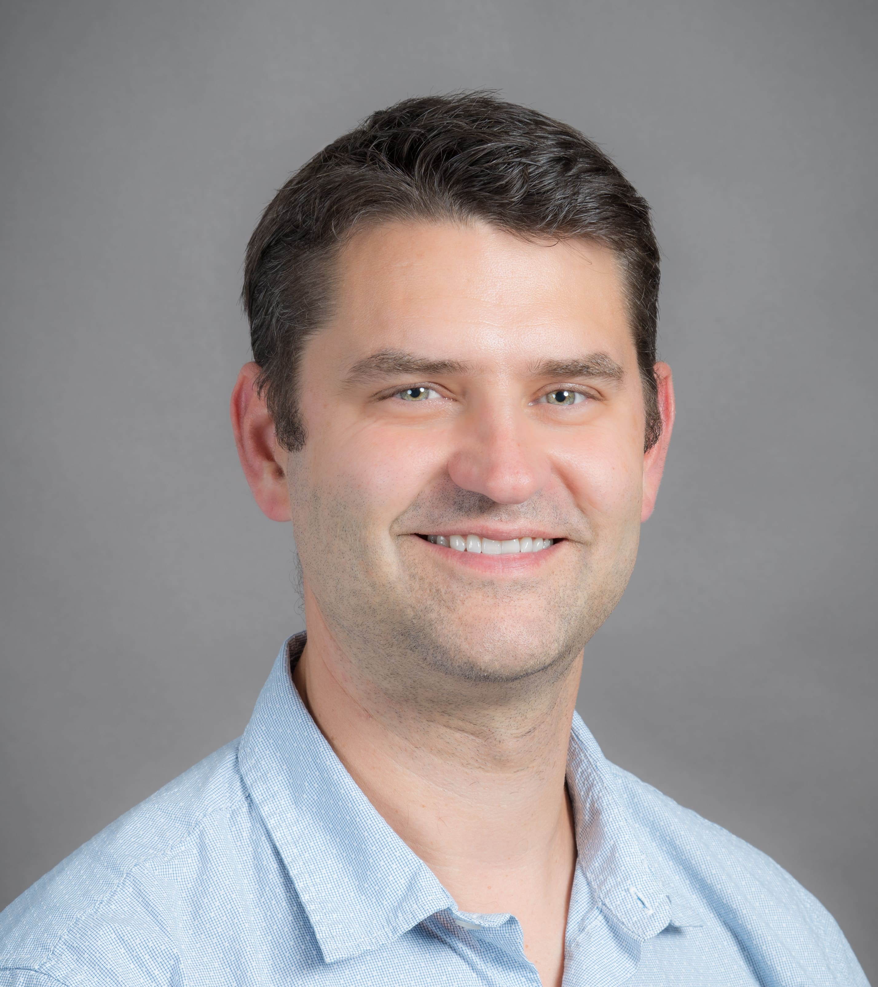 A man with short brown hair and light skin smiles at the camera. He is wearing a light blue collared shirt and is posed in front of a plain gray background.