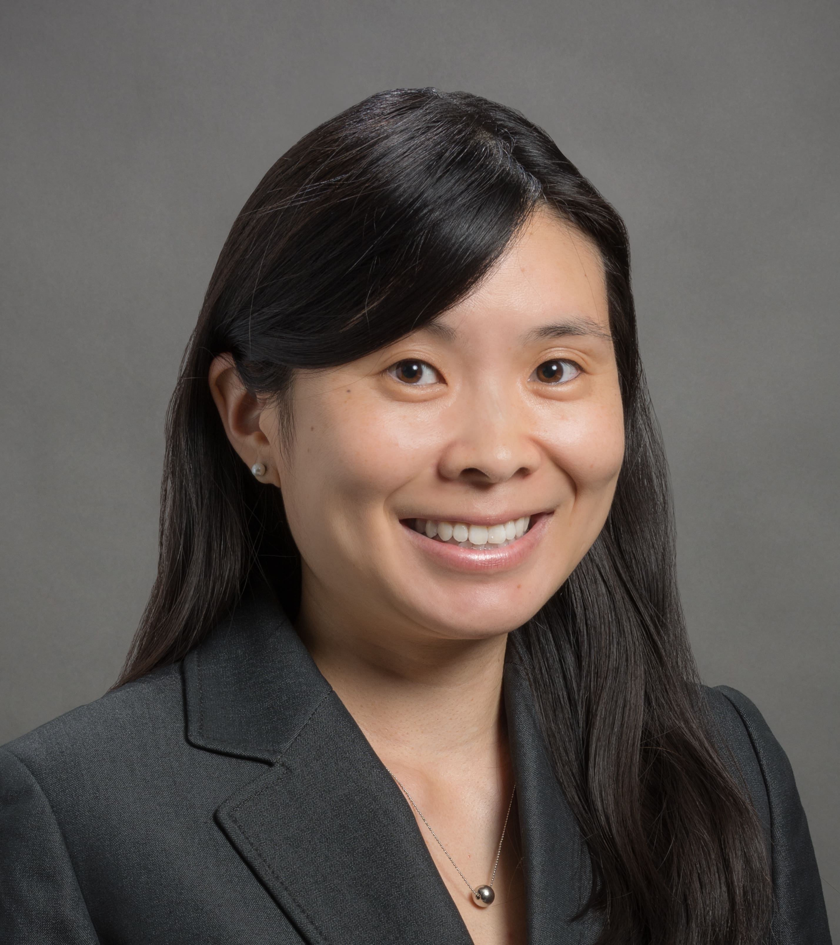 A woman with long black hair, wearing a dark blazer and a necklace, smiles in front of a plain gray background.