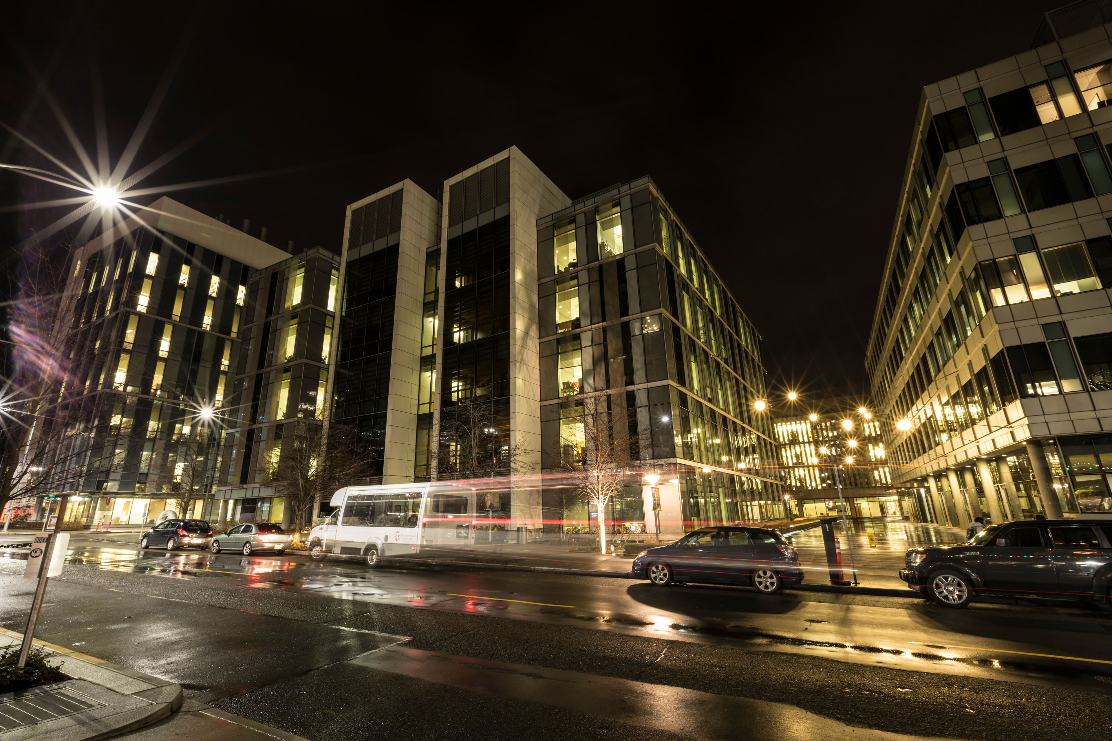 A city street at night with modern office buildings illuminated, cars parked and driving by, and reflections of lights on the wet pavement.