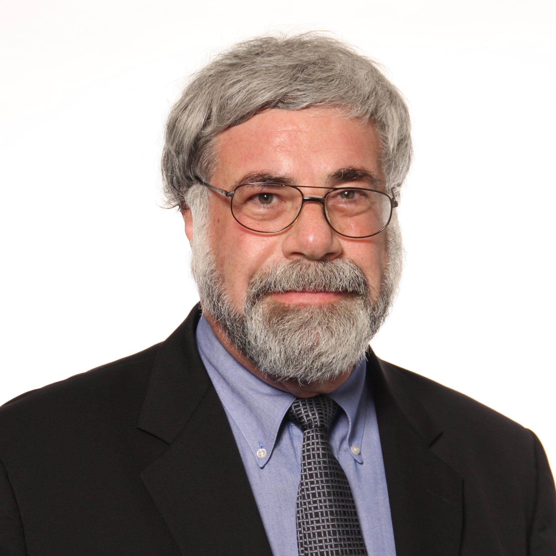 An older man with gray hair, a beard, and glasses wearing a black suit, blue shirt, and patterned tie poses against a plain white background.