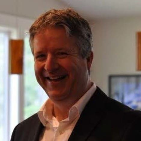 A man with short, graying hair smiles warmly while wearing a dark suit jacket and white shirt in a softly lit indoor setting.