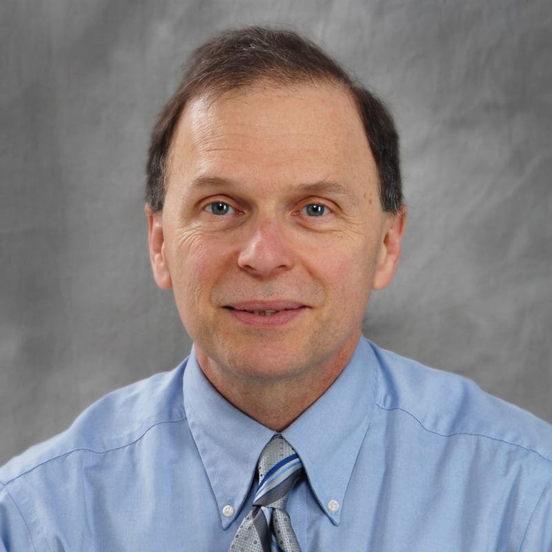 A middle-aged man with short brown hair wearing a light blue dress shirt and striped tie, posing in front of a gray background.