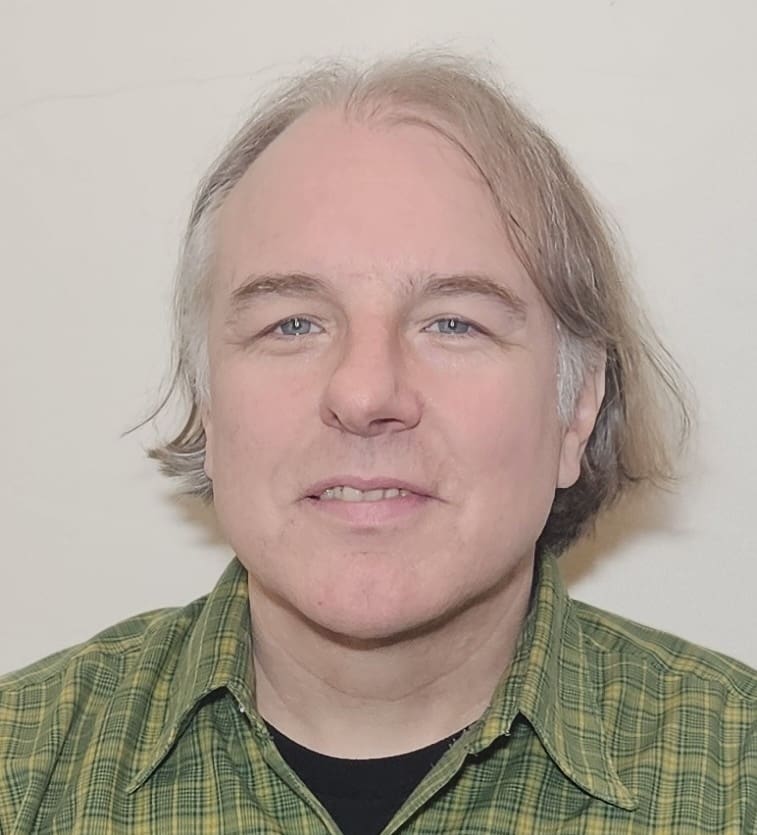 A middle-aged man with light skin, blue eyes, and graying blond hair wears a green plaid shirt and smiles slightly while looking at the camera against a plain light background.