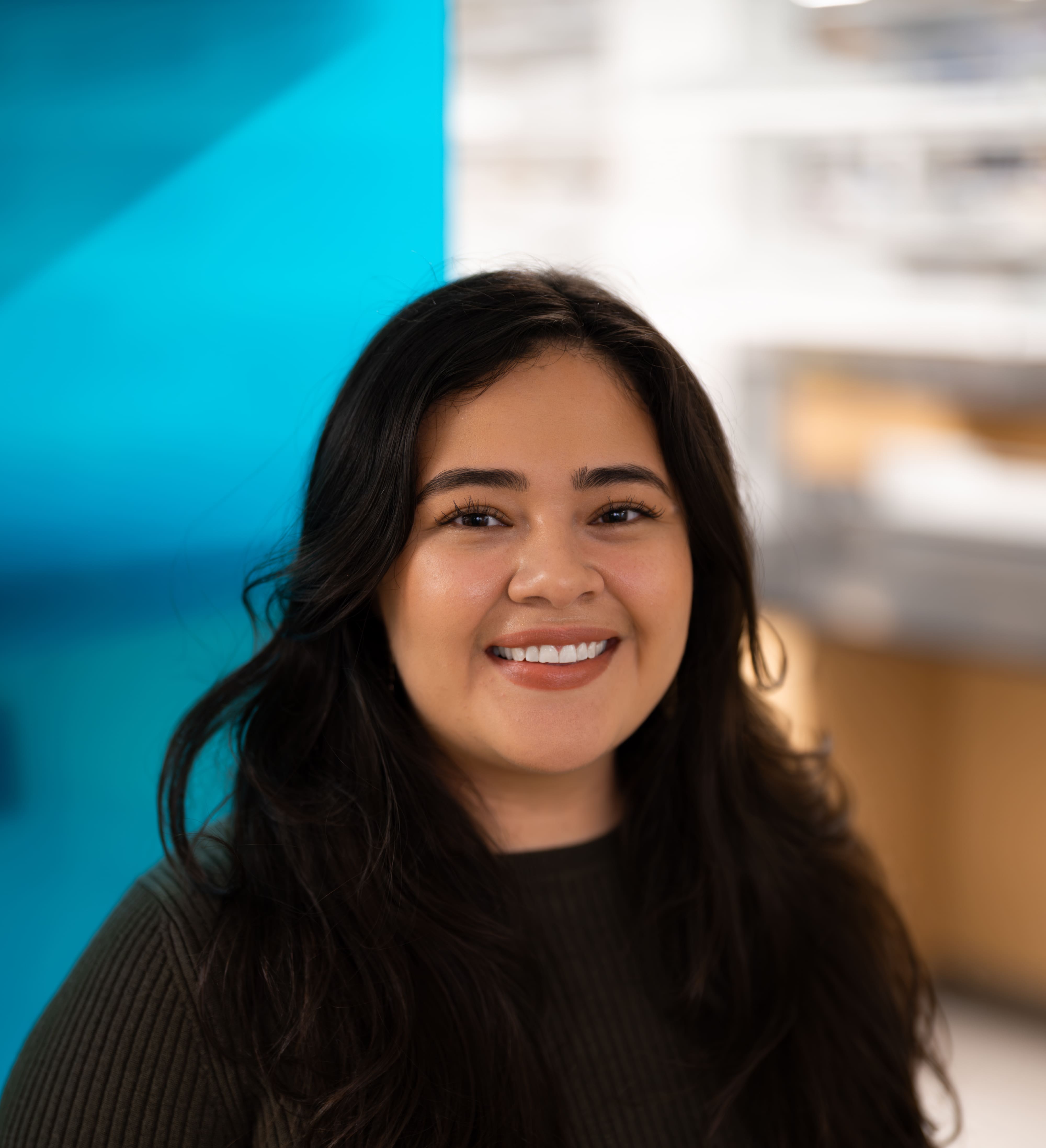 A woman with long dark hair and a green sweater smiles at the camera. The background is blurred, featuring a blue wall and light-colored shelves or cabinets.