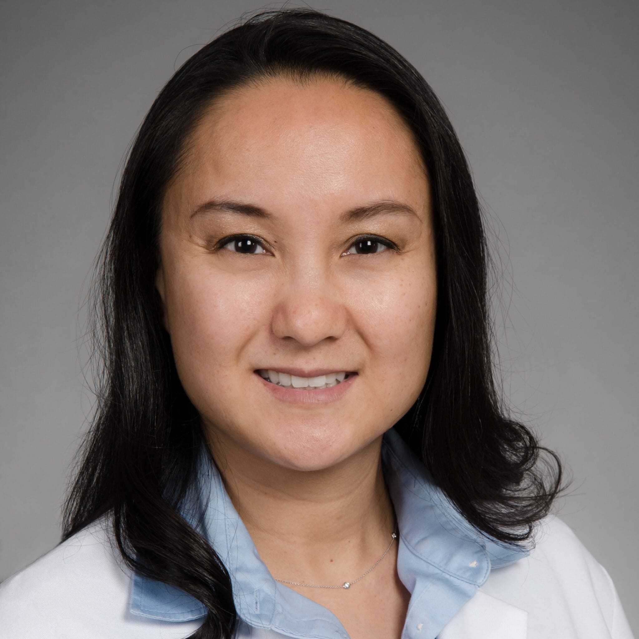 A woman with long dark hair smiles at the camera. She is wearing a white lab coat over a light blue collared shirt, and is posed in front of a plain, light gray background.