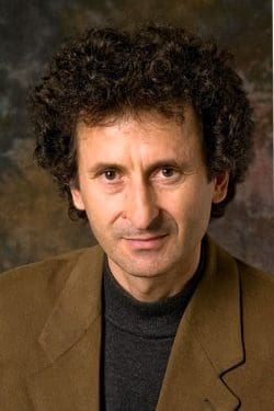 A man with curly dark hair wears a brown blazer over a black turtleneck, posing against a mottled brown and dark background.