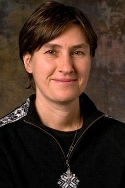 A person with short brown hair is smiling slightly, wearing a black sweater with a white snowflake pattern on the shoulder and a matching snowflake pendant. The background is brown and softly textured.