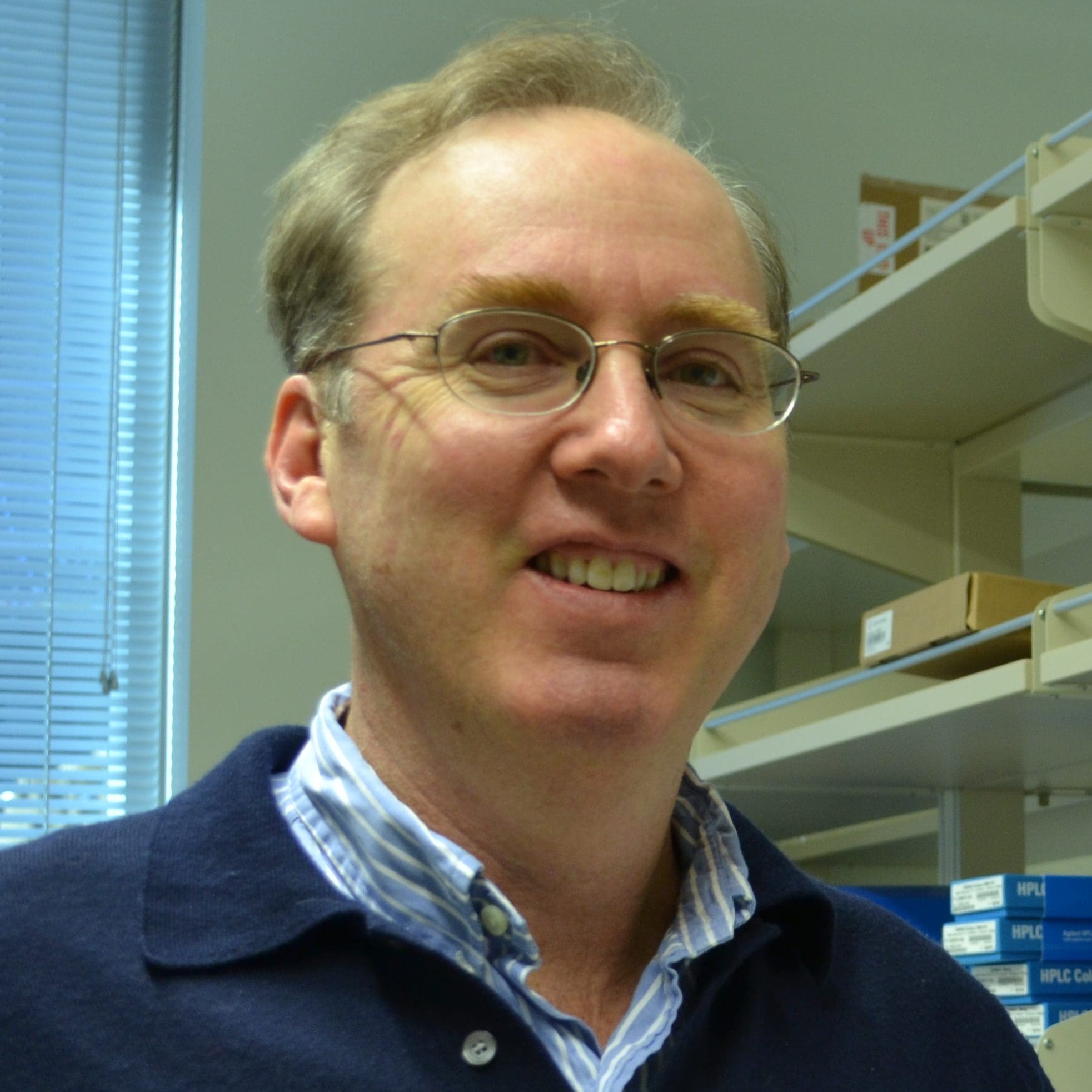 A man wearing glasses and a blue collared shirt stands in an office or laboratory with shelves and boxes in the background, smiling at the camera.