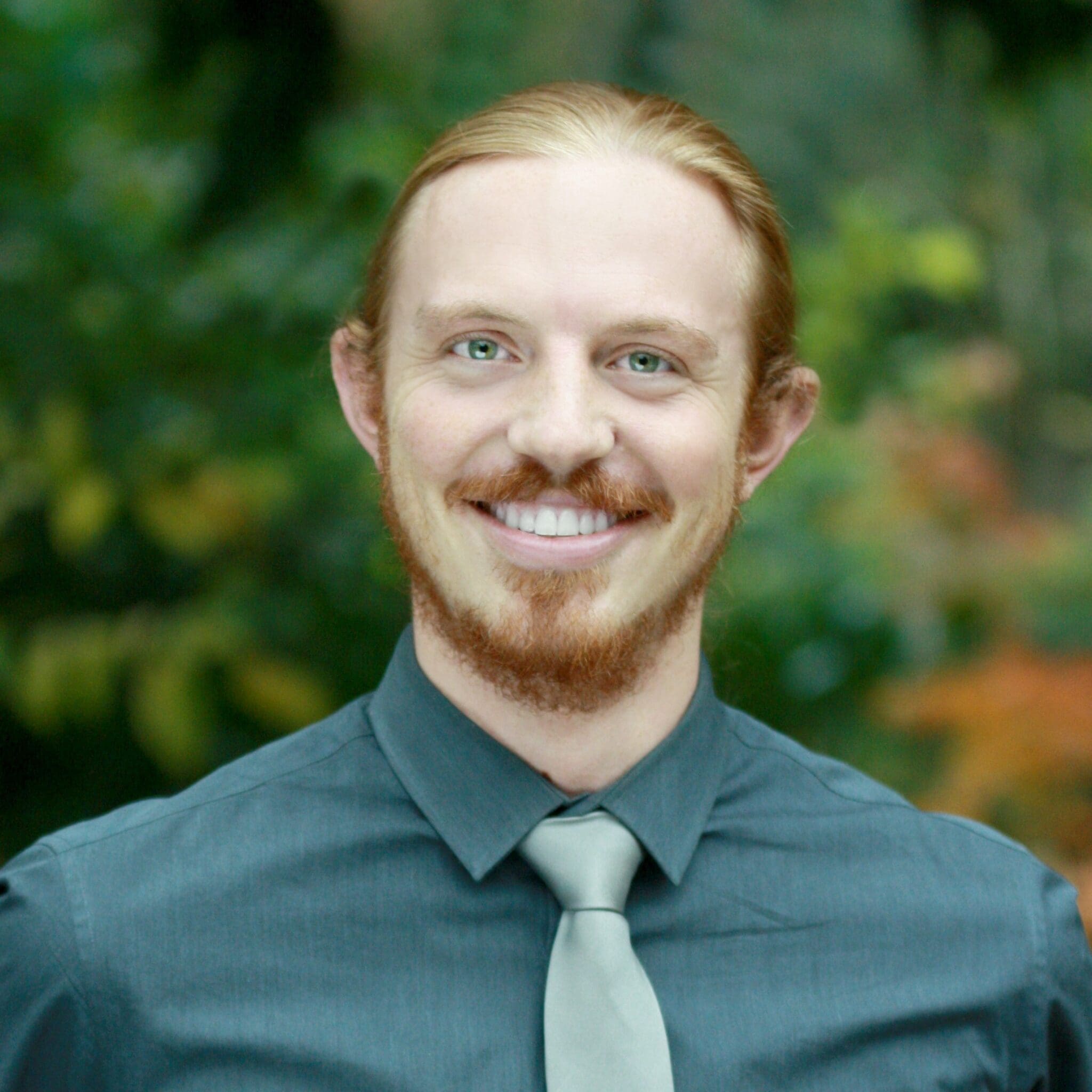 A smiling man with long red hair tied back, a red beard, and mustache, wearing a dark gray shirt and a light gray tie. The background is outdoors with green and yellow blurred foliage.