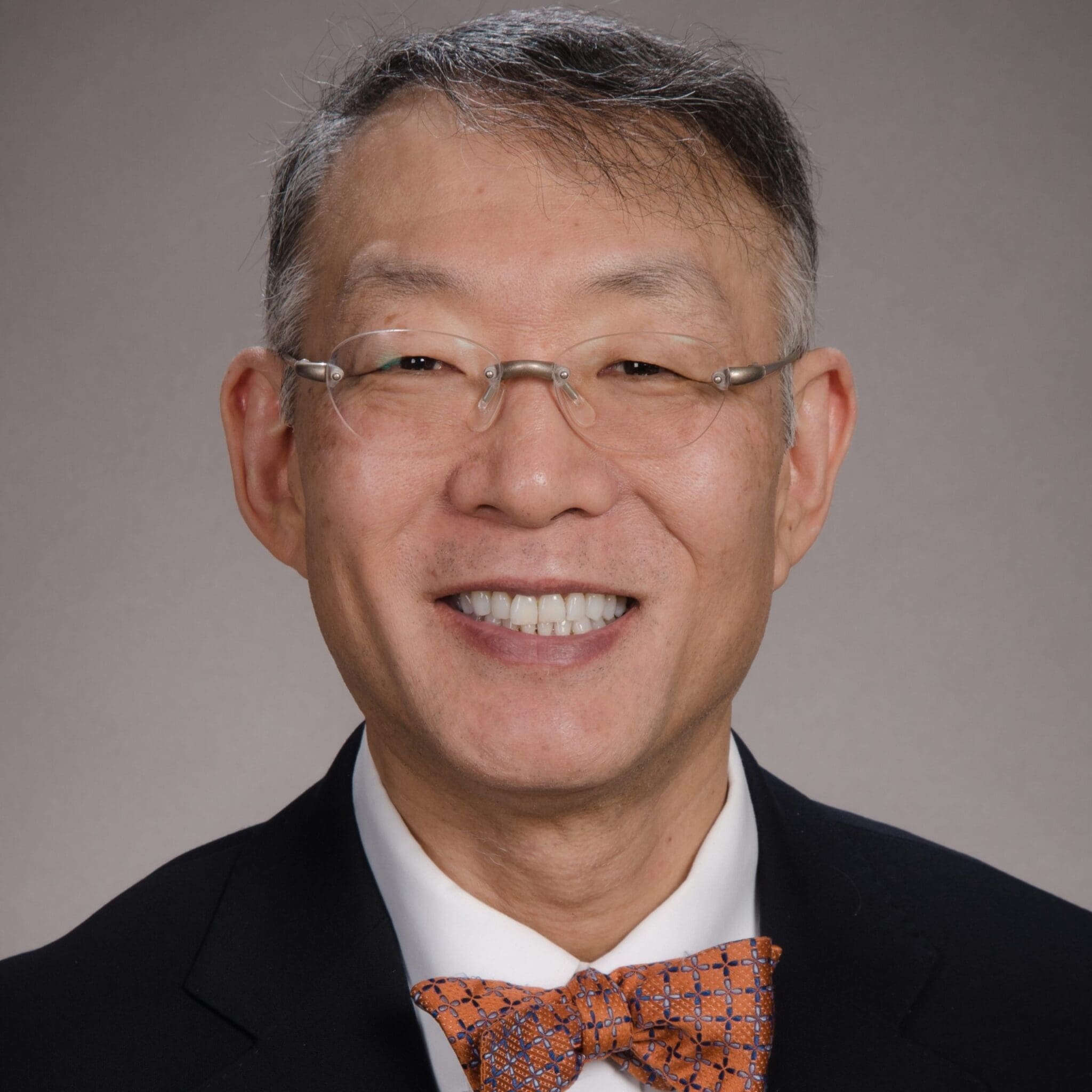 A middle-aged man with glasses smiles at the camera, wearing a black suit, white shirt, and an orange patterned bow tie, with a neutral gray background.