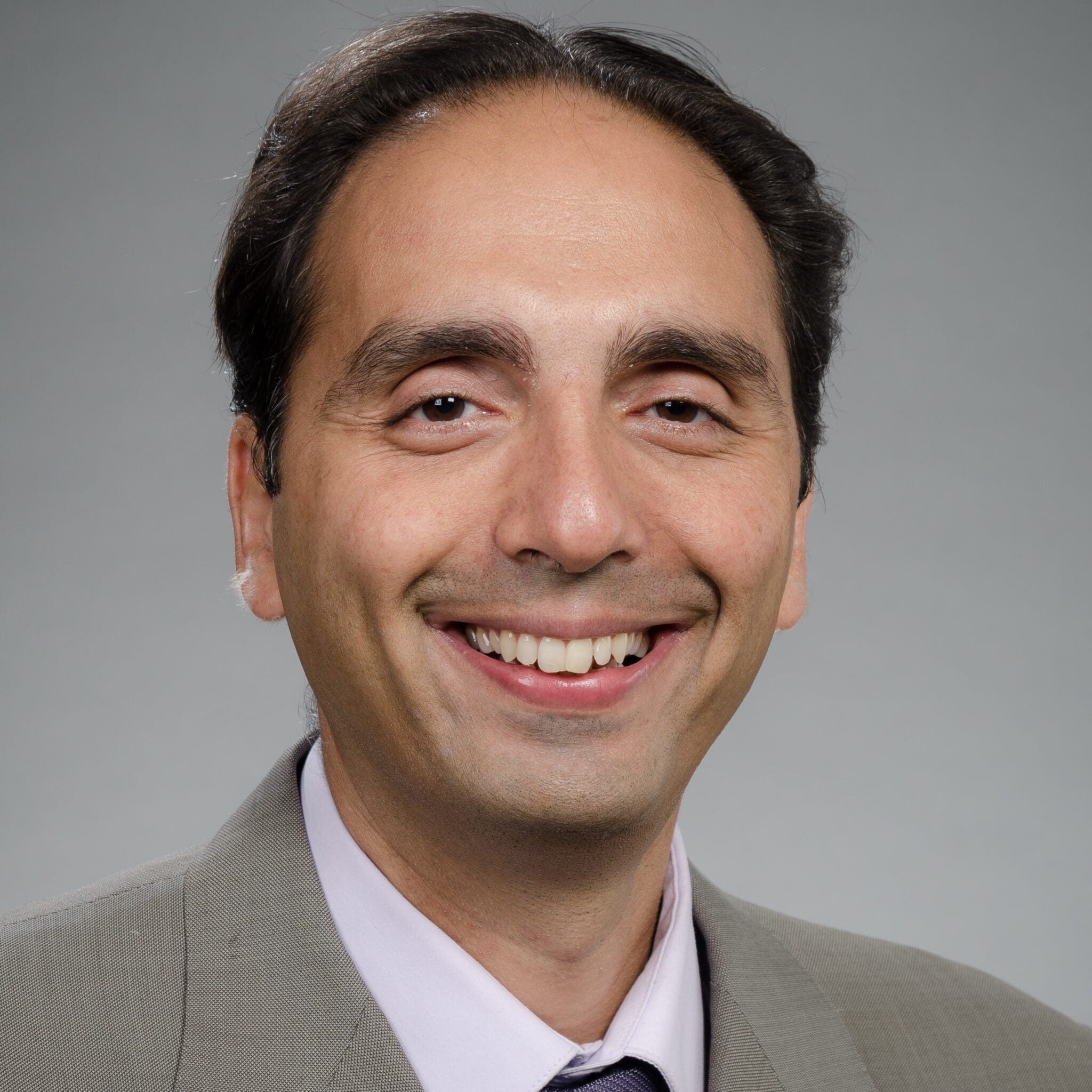 A man with dark hair, wearing a light gray suit, pale lavender shirt, and a dark tie, smiling at the camera against a plain gray background.