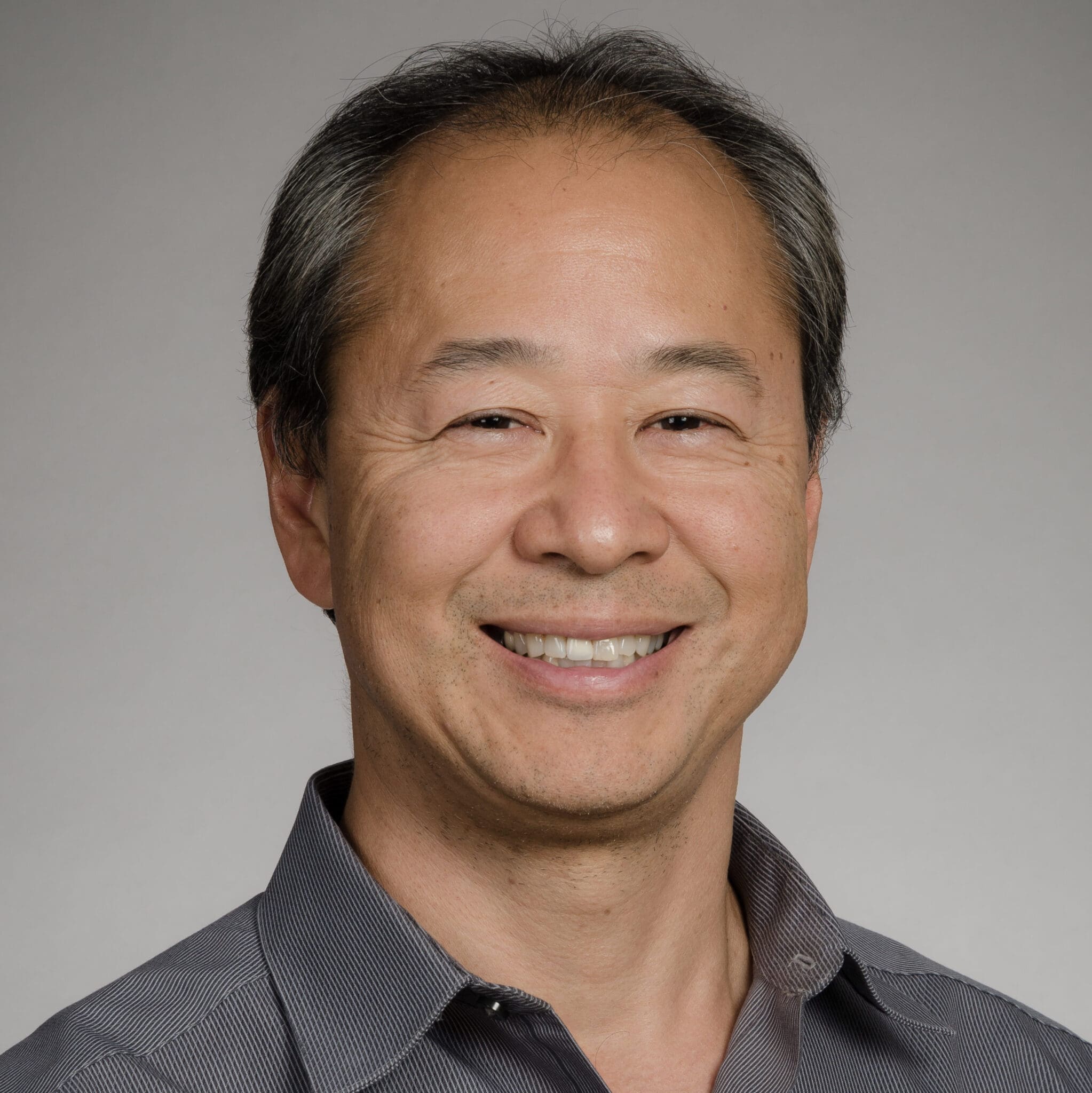 A middle-aged man with short graying hair smiles at the camera. He is wearing a dark collared shirt and is posed in front of a plain light gray background.