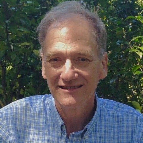 A smiling older man with light skin and short gray hair wears a blue plaid shirt. He is outdoors with green leafy bushes in the background.