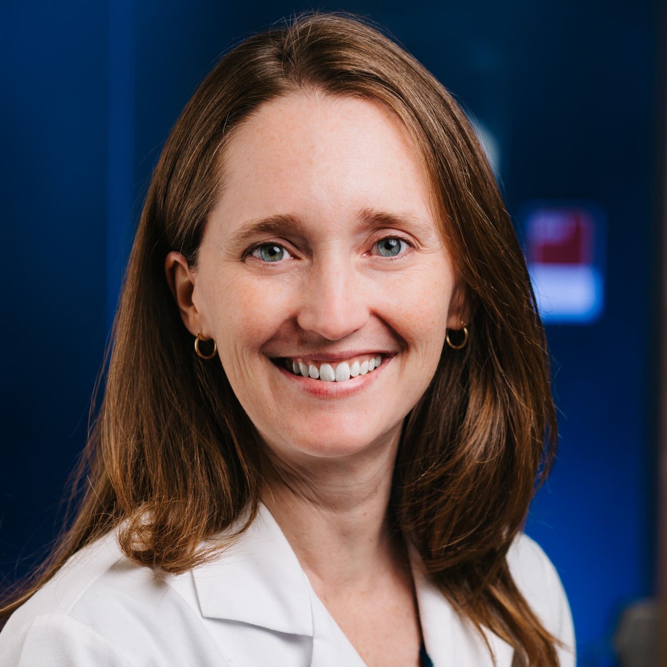 A woman with long brown hair, wearing a white lab coat and small hoop earrings, smiles at the camera against a dark blue background.