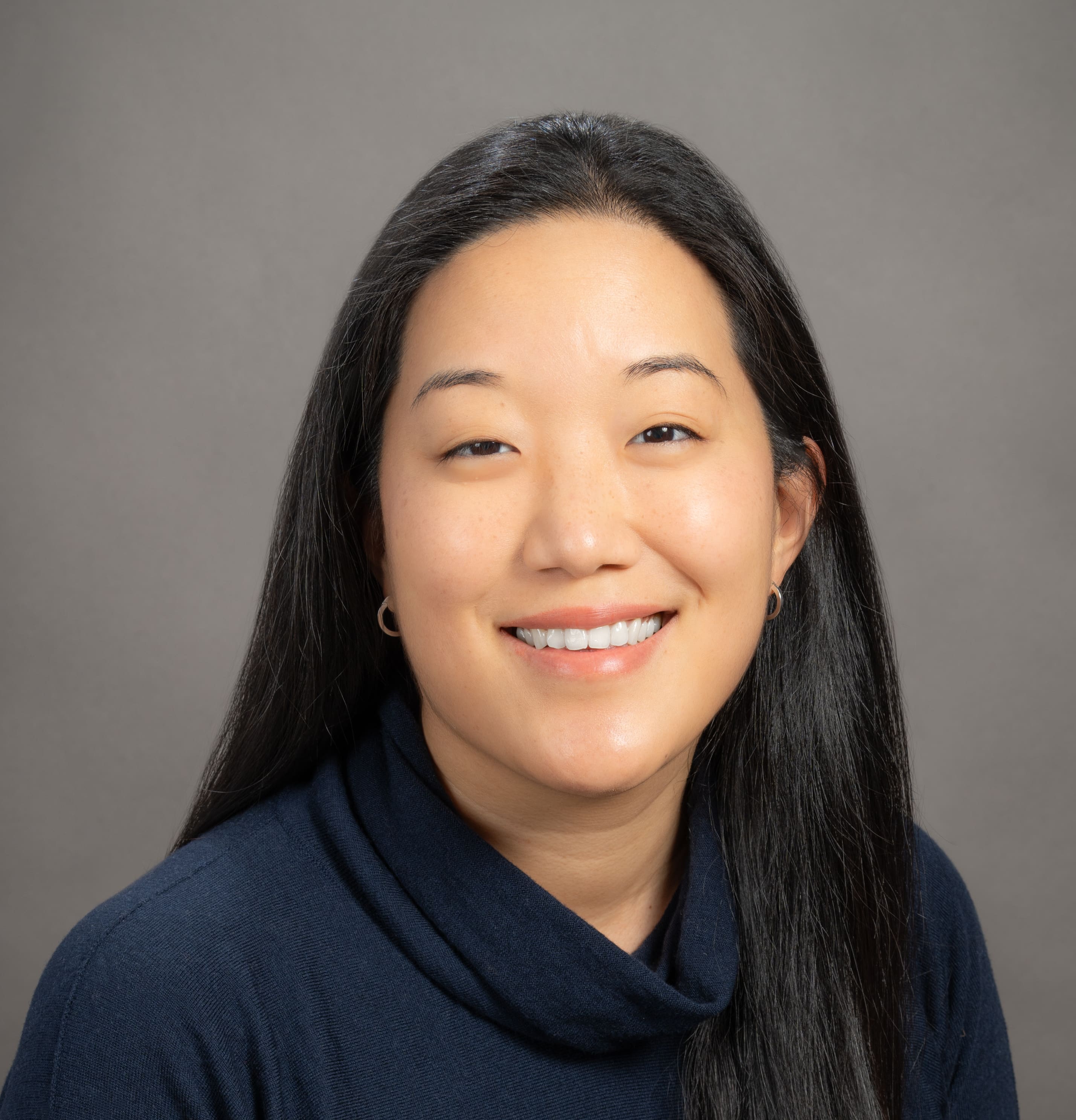 A woman with long straight black hair, wearing a navy blue top, smiles at the camera against a plain gray background.