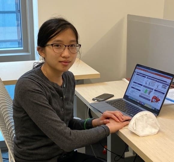 A woman with glasses sits at a desk using a laptop displaying charts. She is wearing a dark sweater and has a white face mask and phone on the desk beside her.