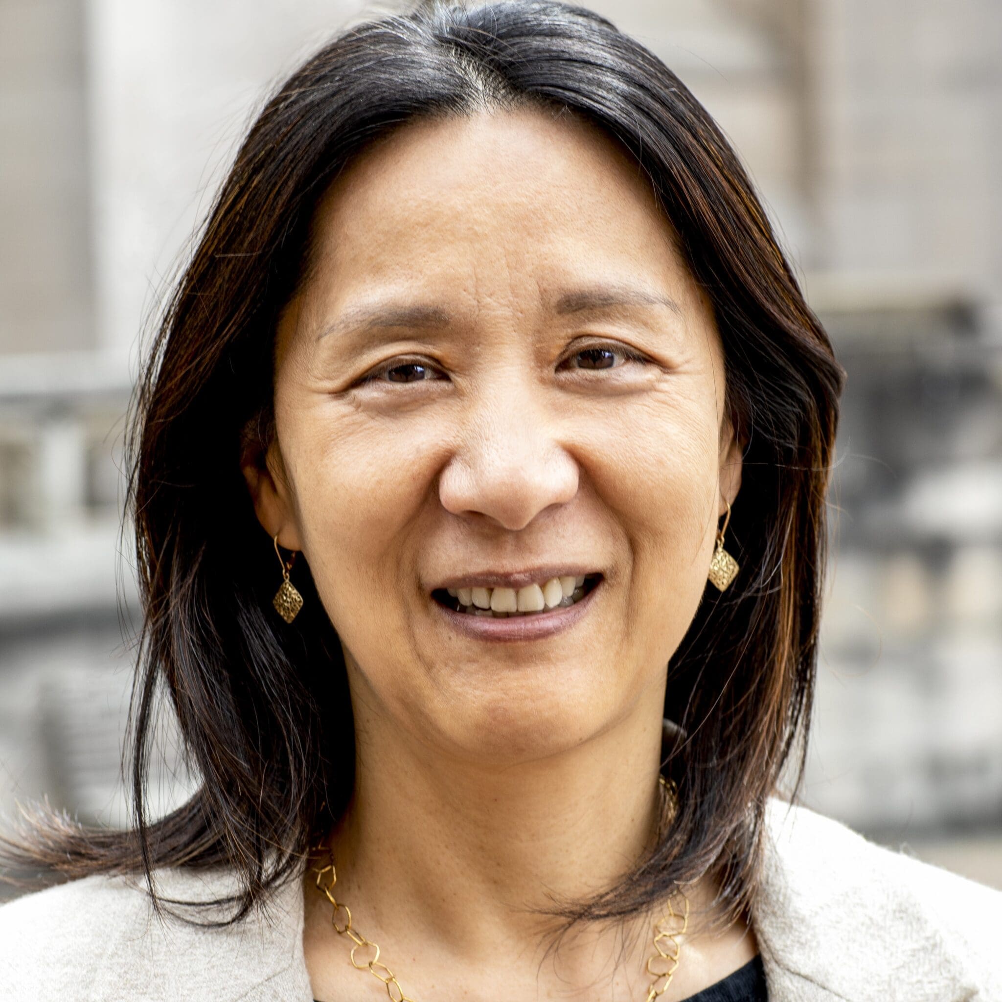 A woman with straight dark hair, wearing gold earrings and a gold necklace, smiles at the camera. She is dressed in a light-colored blazer and standing outdoors with a blurred stone building in the background.