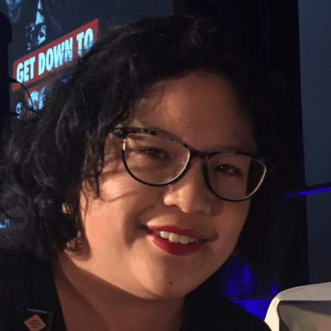 A person with short, wavy black hair and glasses smiles at the camera. The background is dimly lit with a partially visible sign that reads GET DOWN TO.