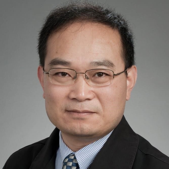 A man wearing glasses, a dark suit jacket, a light blue shirt, and a blue patterned tie poses in front of a plain gray background.